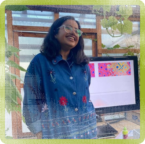 Gaurika Singhal (she/her) - a smiling girl with messy black shoulder length hair in a blue denim embroidered shirt, standing in a greenhouse with a computer screen behind her and beautiful creepers all around