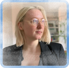Mai Hartmann (she/her), a white woman with bleached blonde hair, blue eyes, glasses and slightly red flushed skin. As instructed by her photographer/partner, her face is turned to the side towards the light and she is flashing a subtle LinkedIn-friendly smile.