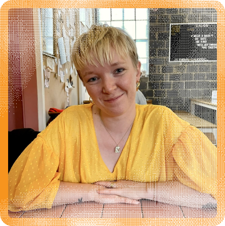 Henriette Friis (she/her), a young white woman with a blonde pixie haircut, wearing a bright yellow dress. She is sitting at a table with pink tiles, and she is resting her arms in front of her. Two tattoos are visible on her arms. She is smiling at the camera with her head slightly tilted.