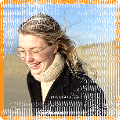 Maja Styrk Andersen (she/her) a person with light skin tone, long hair with light highlighters, and rectangular glasses. She is in the middle of a laugh, looking down. There is a beach background.