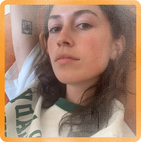 Maya Ellen Hertz (they/them), a white person with brown, curly hair, looks at the camera from a side-angle. Maya wears an oversized white t-shirt with dark green lettering, and sits against an orange couch. Their arm is raised above their head, showing a tattoo on their arm.